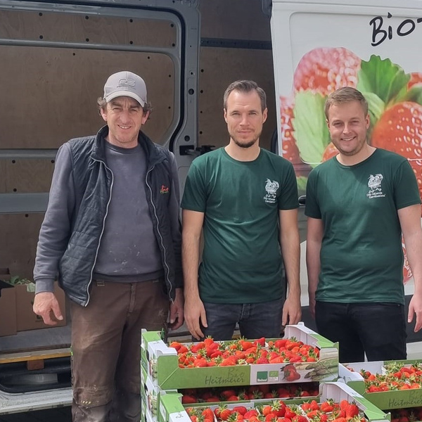 KI generiert: Drei Männer stehen vor einem Lieferwagen mit Erdbeerkisten. Text: "Bio Obsthof".