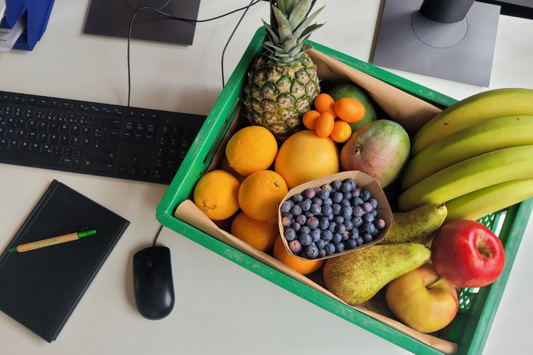KI generiert: Eine Kiste mit verschiedenen Früchten wie Ananas, Bananen, Äpfel, und Blaubeeren auf einem Schreibtisch.