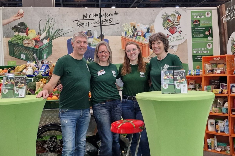 KI generiert: Vier Personen an einem Stand mit Lebensmitteln, Bannertext: "Wir bio liefern die Region seit 1995!"