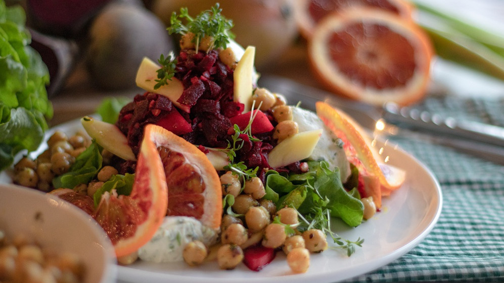 KI generiert: Das Bild zeigt eine Schüssel mit einem bunten Salat aus Rote-Bete-Scheiben, Karottenstreifen, Feldsalat, gehackten Nüssen und einem Klecks Sahne oder Joghurt in der Mitte. Der Salat liegt auf einem Holztisch.