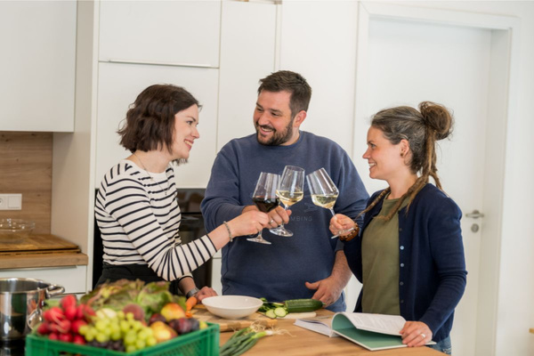 KI generiert: Das Bild zeigt zwei Personen in grüner Arbeitskleidung, die lächelnd eine Kiste mit frischem Obst und Gemüse halten. Im Hintergrund sind weitere Kisten mit Tomaten, Äpfeln und Salat zu sehen, was auf ein Lager oder eine Verkaufsstelle für frische Lebensmittel hinweist.