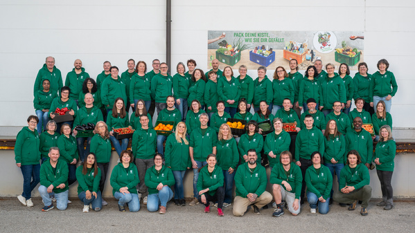 KI generiert: Eine große Gruppe in grünen Jacken posiert mit Gemüse. Text: "PACK DEINE KISTE, WIE SIE DIR GEFÄLLT."
