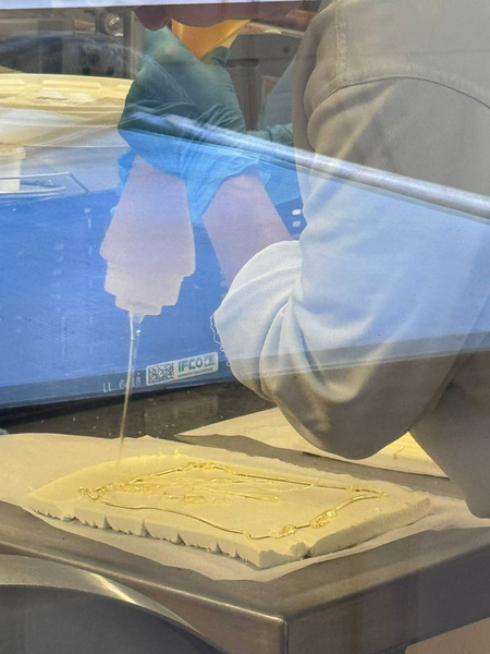 KI generiert: Person mit Handschuhen trägt Flüssigkeit aus Flasche auf rechteckigen Teig auf.